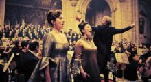 Two women sing as a man conducts a full orchestra behind them in a church.