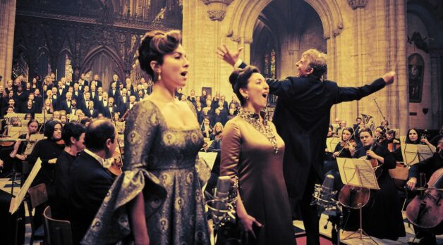 Two women sing as a man conducts a full orchestra behind them in a church.