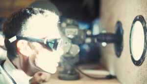 A man in lab glasses stands in front of a porthole, through which a blinding light shines.
