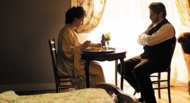 A woman eats at a small wooden table while a man sits opposite looking at her.