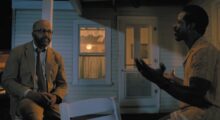 Two black men sit and talk on a porch at night.