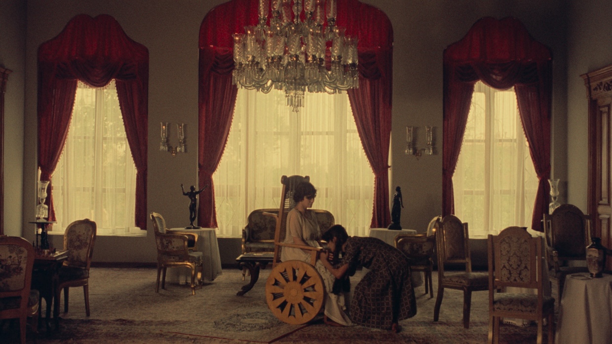 A woman kneels before in a woman in a wheelchair in an opulent room.
