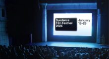 A crowd sits in a dark theater, the "Sundance Film Festival 2024" logo illuminates the screen.