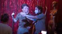 On a stage with red sequins at the back, a man in a surgical mask is gesturing in front of a man who is has a disfigured face.