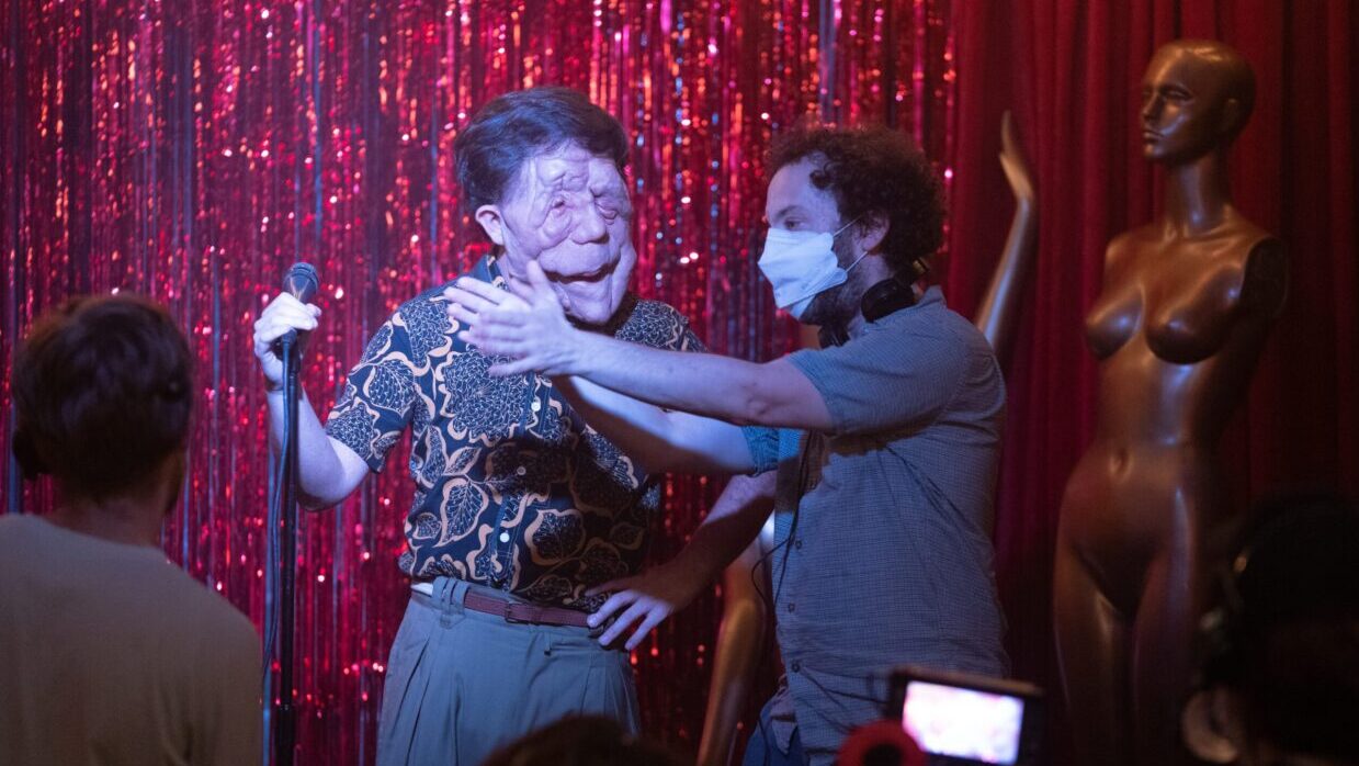 On a stage with red sequins at the back, a man in a surgical mask is gesturing in front of a man who is has a disfigured face.