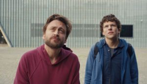 Two white men, one wearing a red hoodie and one wearing a blue zip-up, gaze upward.