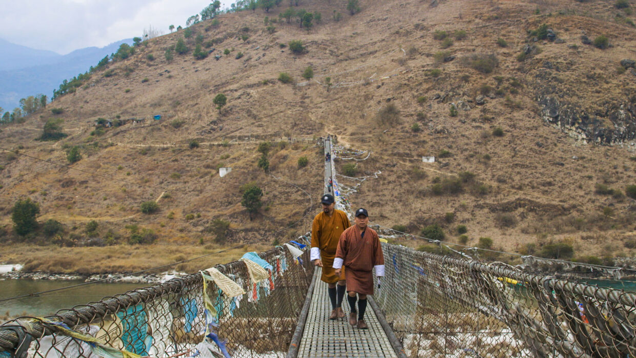 Two figures cross a bridge in an Alpine landsdcape in Bhutan.