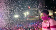 A Filipina woman wearing a pink striped polo and mask stands amid a crowd as rainbow confetti falls.