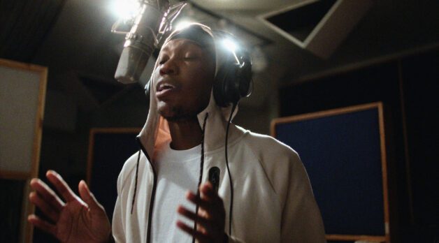 A young Black man wearing a white t-shirt and hoodie sings into a microphone at a recording studio.