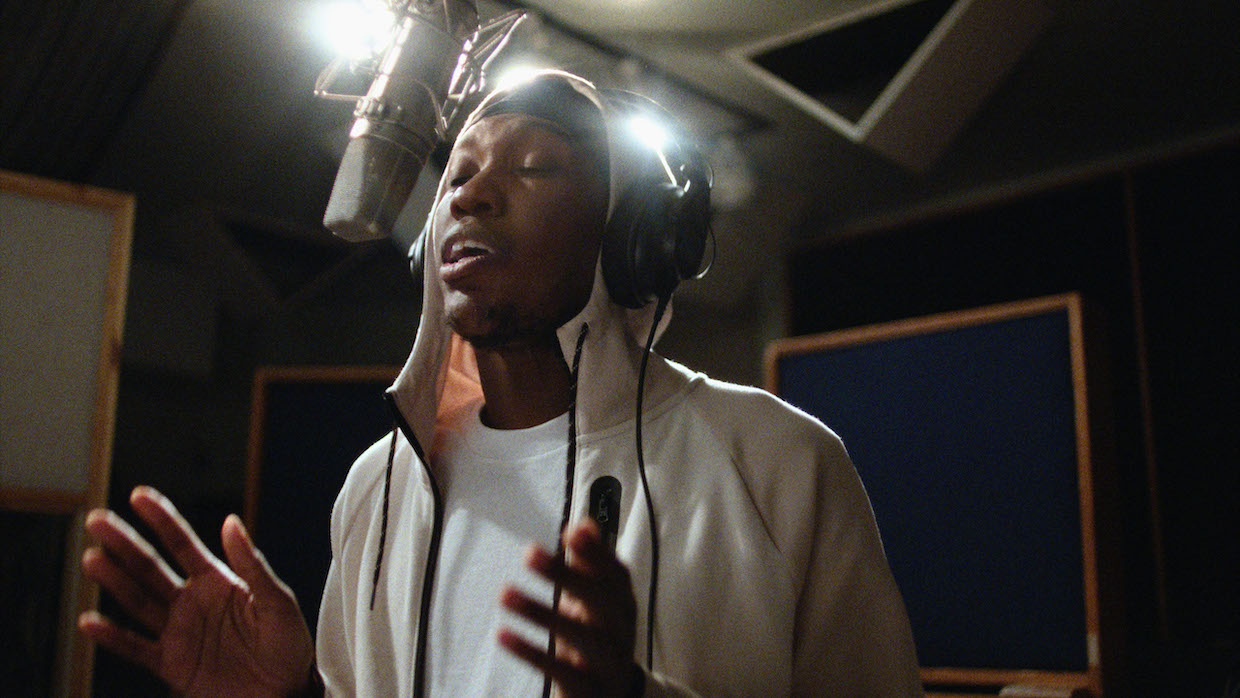 A young Black man wearing a white t-shirt and hoodie sings into a microphone at a recording studio.