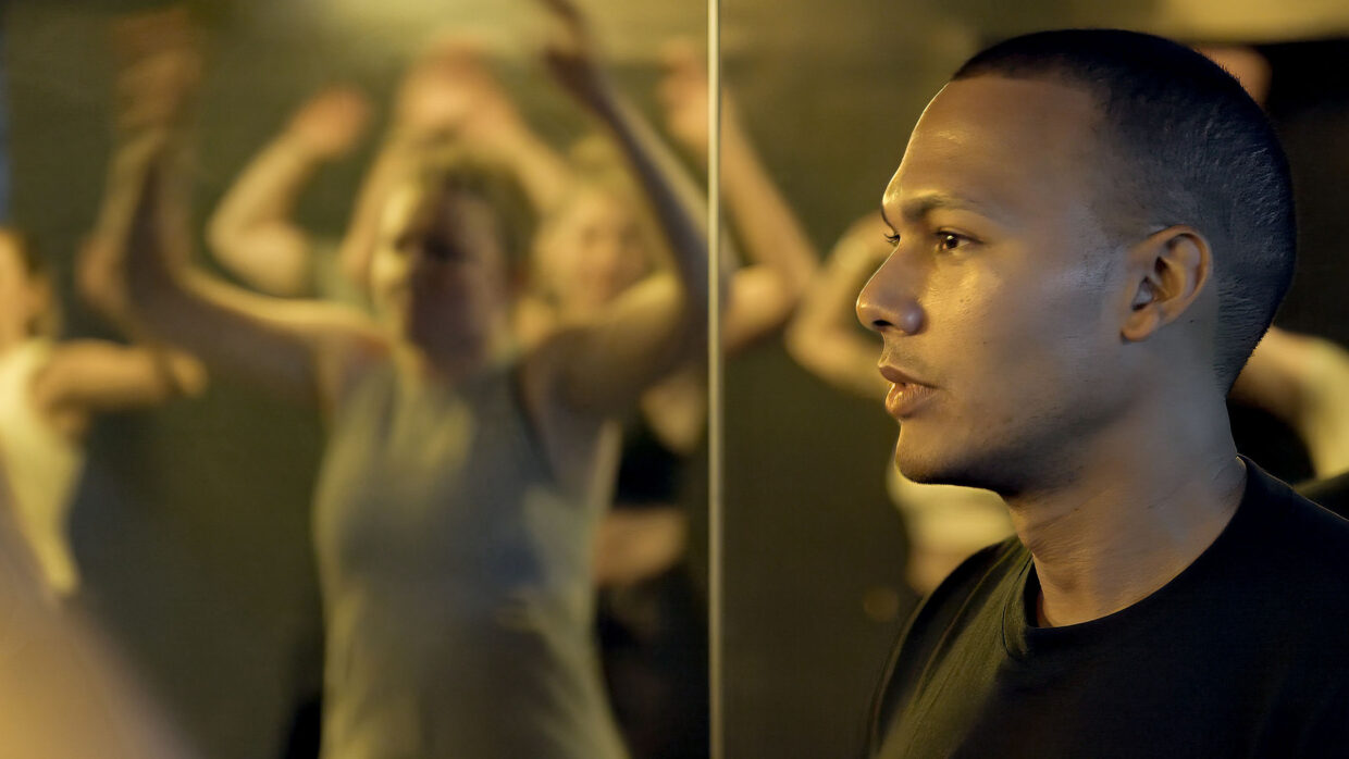 A Hispanic man looks to the left in the foreground, while in the background a woman is seen flexing in a mirror.