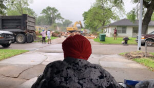 An obscured person wearing a gray coat and red headwear looks across a street toward a backhoe and wooden planks.