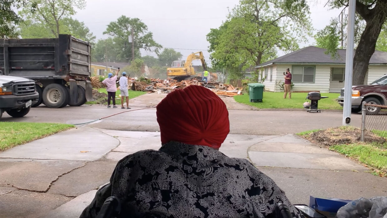 An obscured person wearing a gray coat and red headwear looks across a street toward a backhoe and wooden planks.