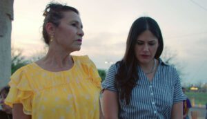 Two women stand outside during twilight. On the left, an older woman wears a ruffled yellow shirt and looks to her left. On the right, a younger woman wears a blue and white striped blouse and looks downward.