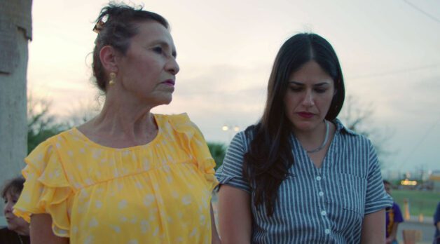Two women stand outside during twilight. On the left, an older woman wears a ruffled yellow shirt and looks to her left. On the right, a younger woman wears a blue and white striped blouse and looks downward.