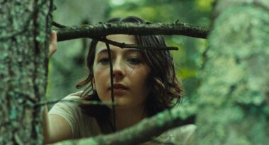 A young woman with wavy brown hair looks out from behind a tree stump and branches.