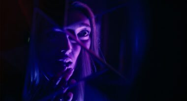 A woman bathed in purple light looks into a broken mirror