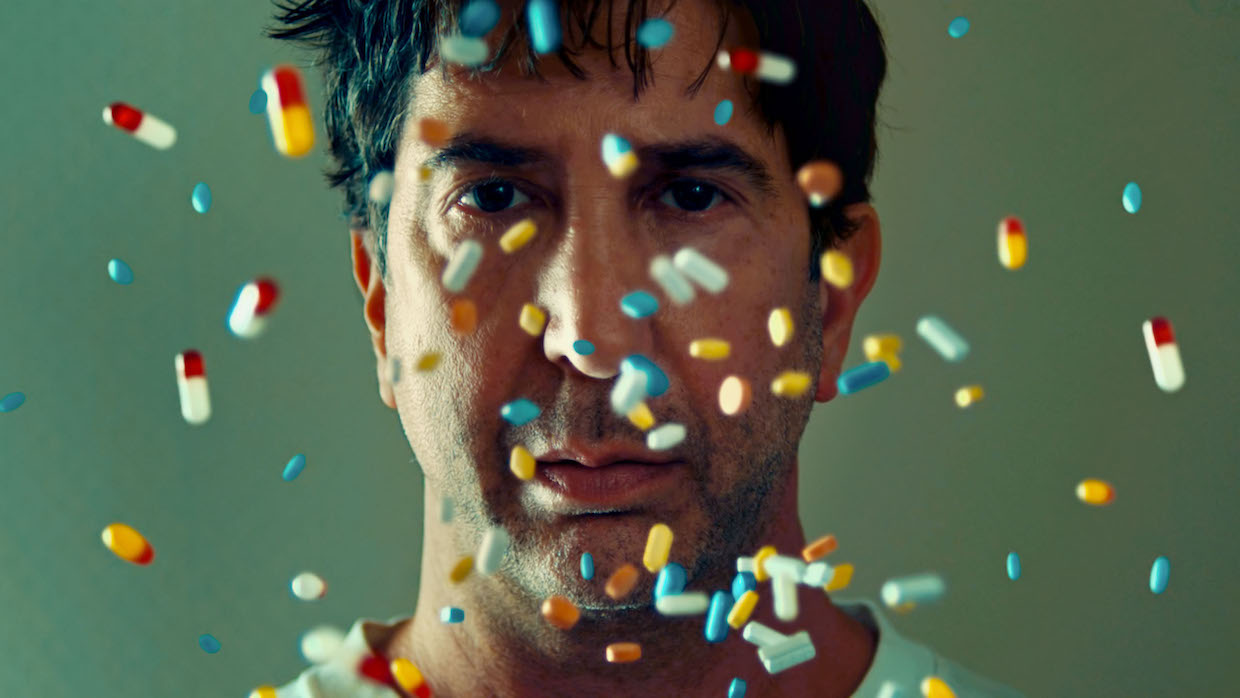 A shot of David Schwimmer's face as colorful prescription pills fall in front of it.