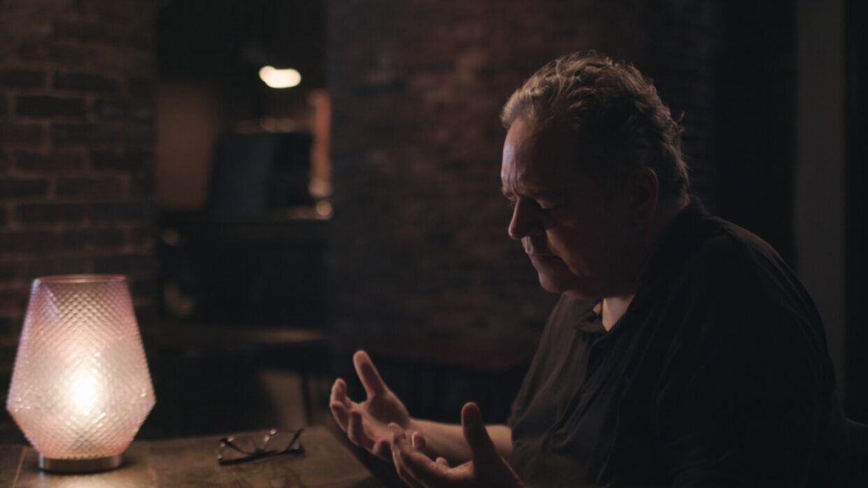 A white man stares at his hands in a dimly lit room with brick walls.