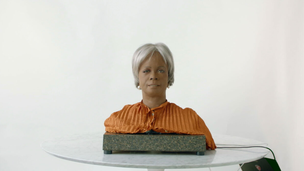 BINA48, a humanoid AI consisting of shoulders and head with dark skin and gray hair, is sitting on a table.