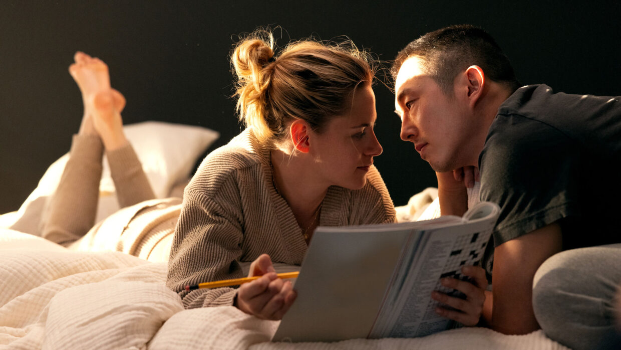 On a white bedspread, a white woman holding a magazine and an Asian man look into one another's eyes.