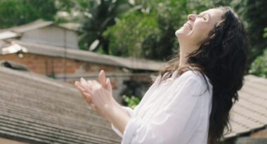 A white woman with long, curly dark hair wears a white tunic and stands smiling in the sunlight.