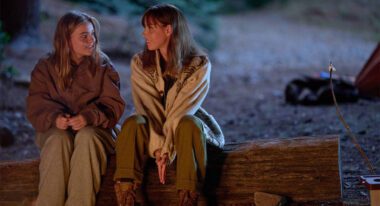 Two women, one younger and one older, sit on a log in the forest illuminated by a campfire.