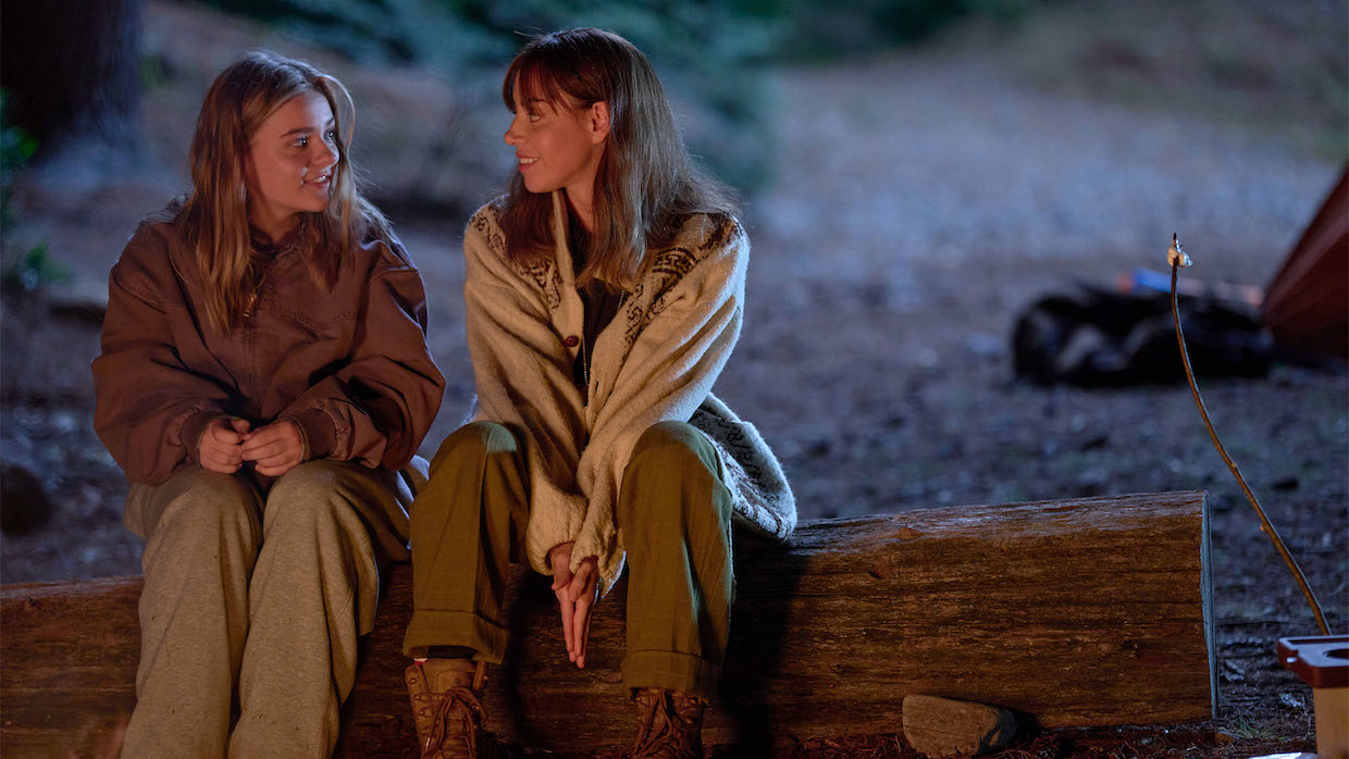 Two women, one younger and one older, sit on a log in the forest illuminated by a campfire.