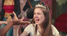 A young girl wearing a fake crown smiles while sitting in a wheelchair.