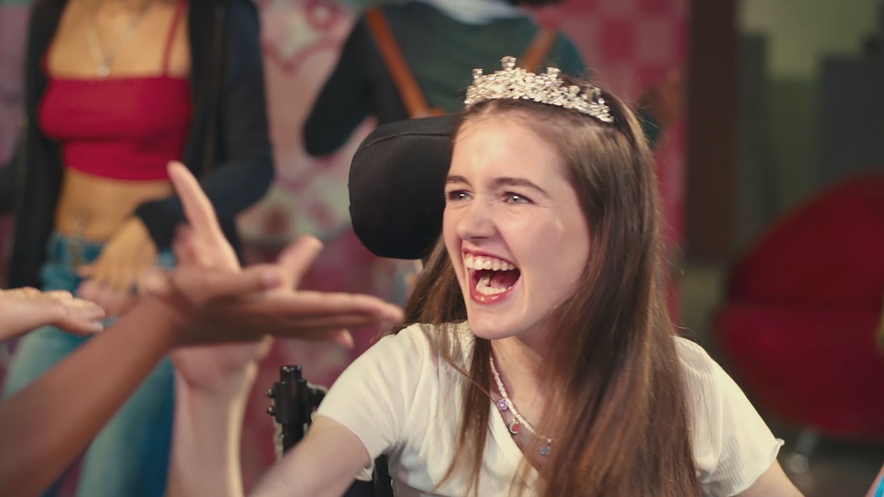 A young girl wearing a fake crown smiles while sitting in a wheelchair.