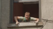 A white man with wet hair leans out of a window on a bright day.