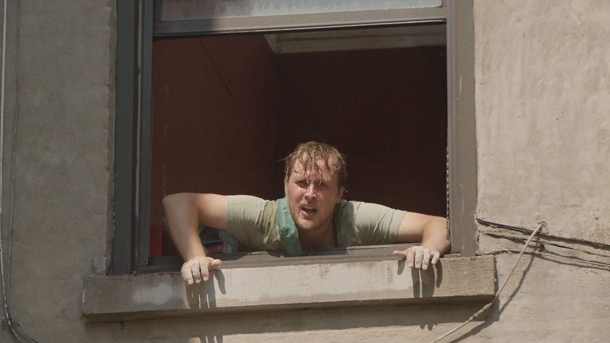 A white man with wet hair leans out of a window on a bright day.