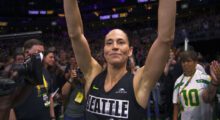 Basketball player Sue Bird raises her arms in victory and smiles at the camera.