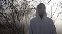 A young man in a white hoodie is looking toward the camera. There are bare trees behind him.
