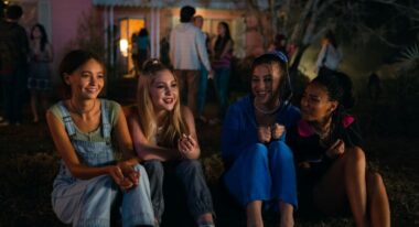 In the foreground, a group of adolescent girls smile. In the background, a group of adults are talking to one another.