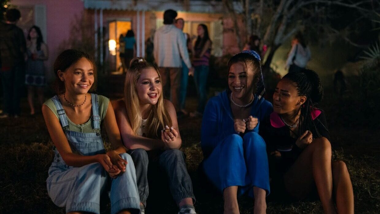 In the foreground, a group of adolescent girls smile. In the background, a group of adults are talking to one another.