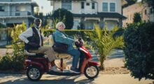 An elderly Black man and an elderly white woman are sitting on a red electric scooter.