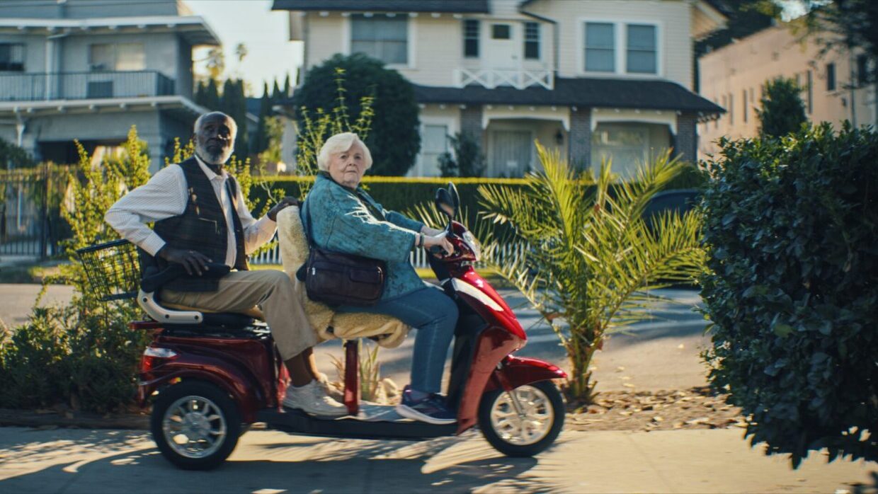An elderly Black man and an elderly white woman are sitting on a red electric scooter.