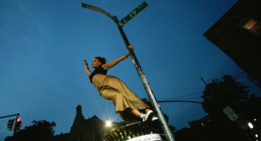 A young Black woman wearing yellow pants and a crop top hangs off of a street post.