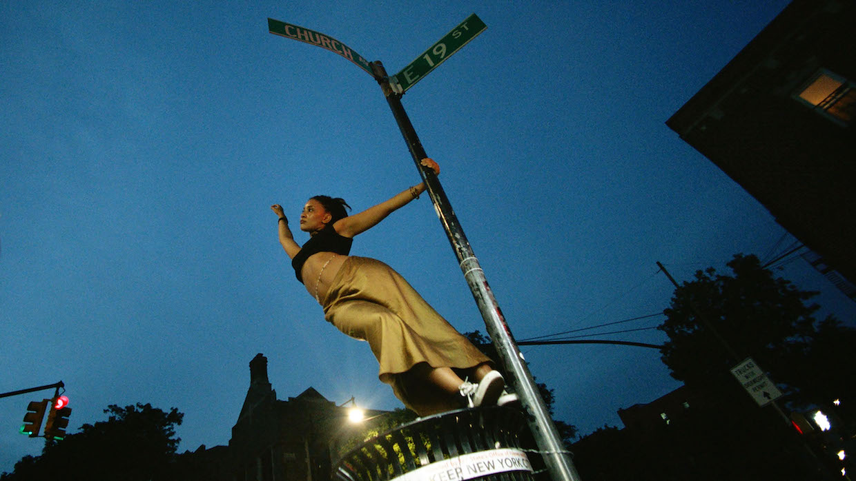 A young Black woman wearing yellow pants and a crop top hangs off of a street post.