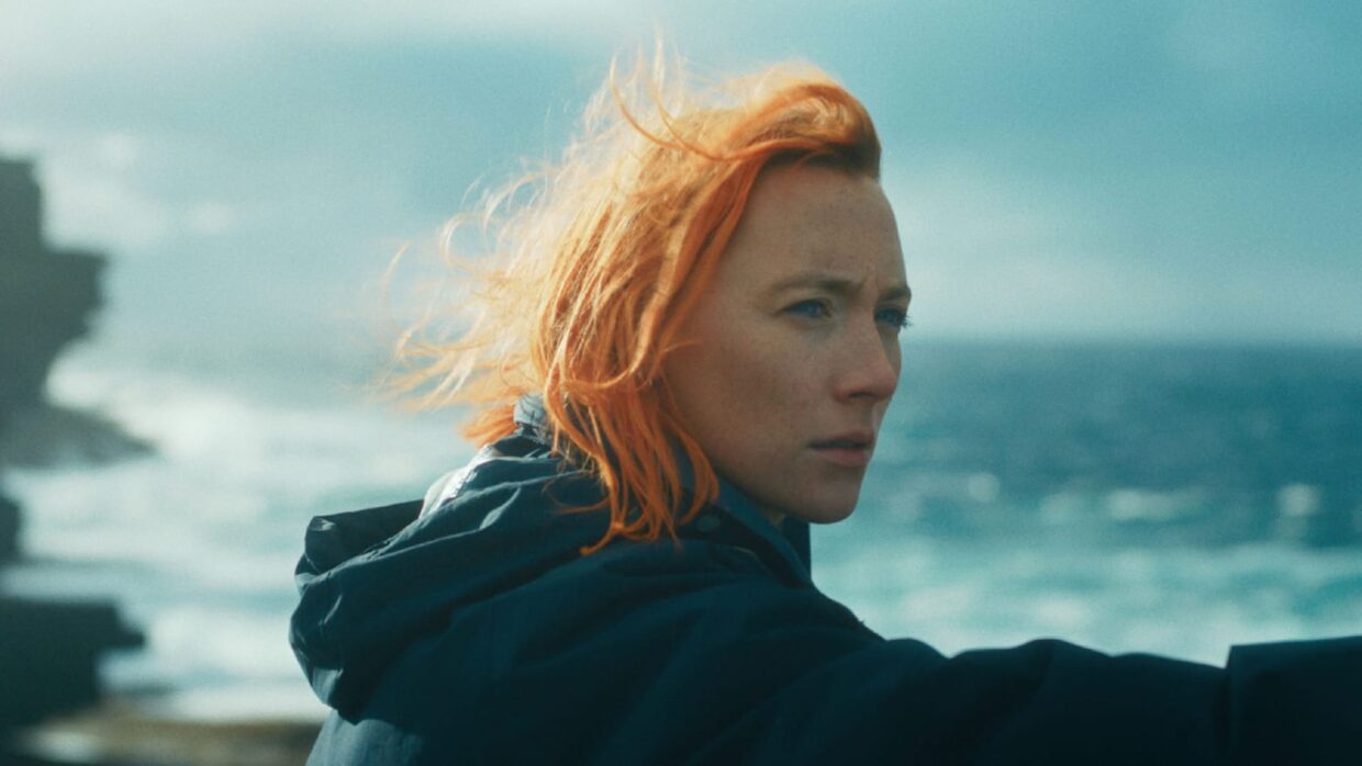 A young woman played by actress Saoirse Ronan with orange hair wearing a black hoodie standing against the sea