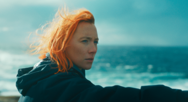 A red-headed woman is looking out off-camera to the right with the ocean visible behind her.