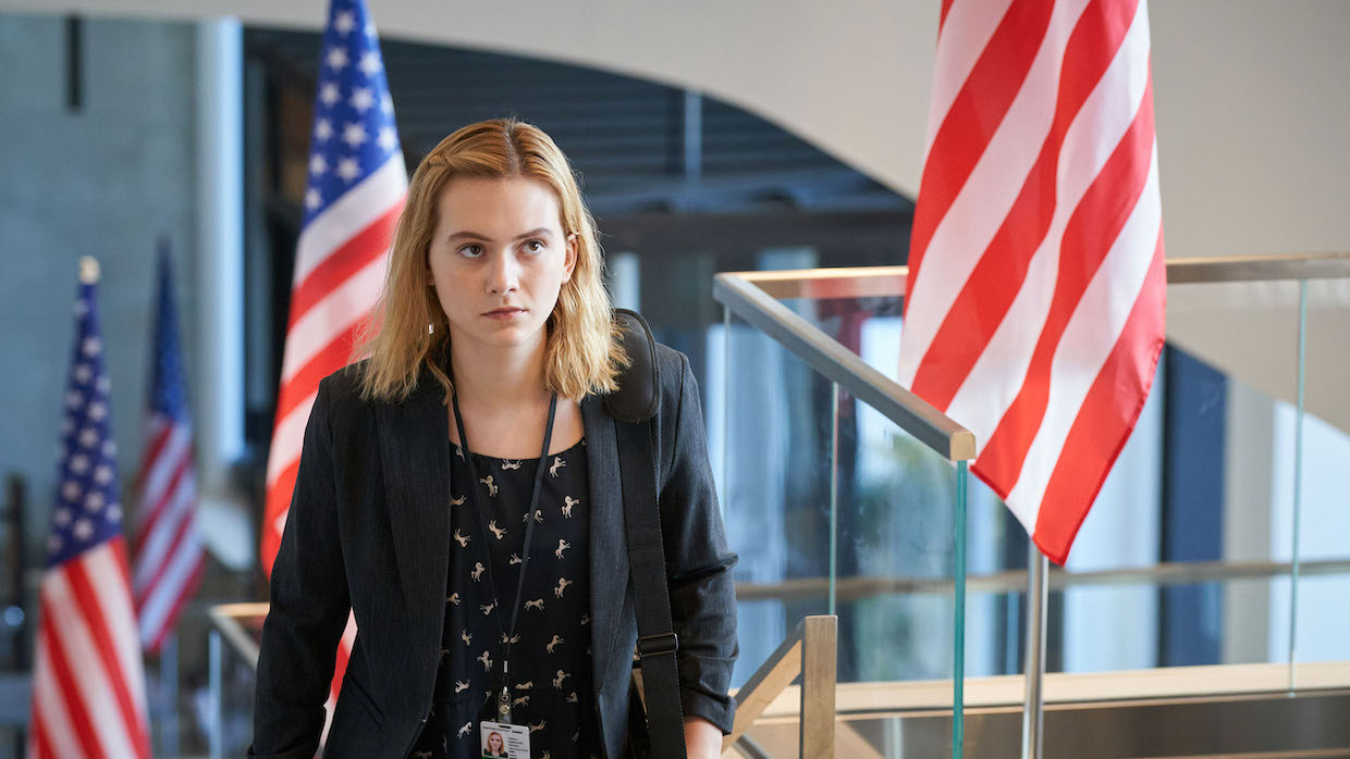 A young woman with blond hear wears a blazer and stands in a building lobby adorned with American flags.