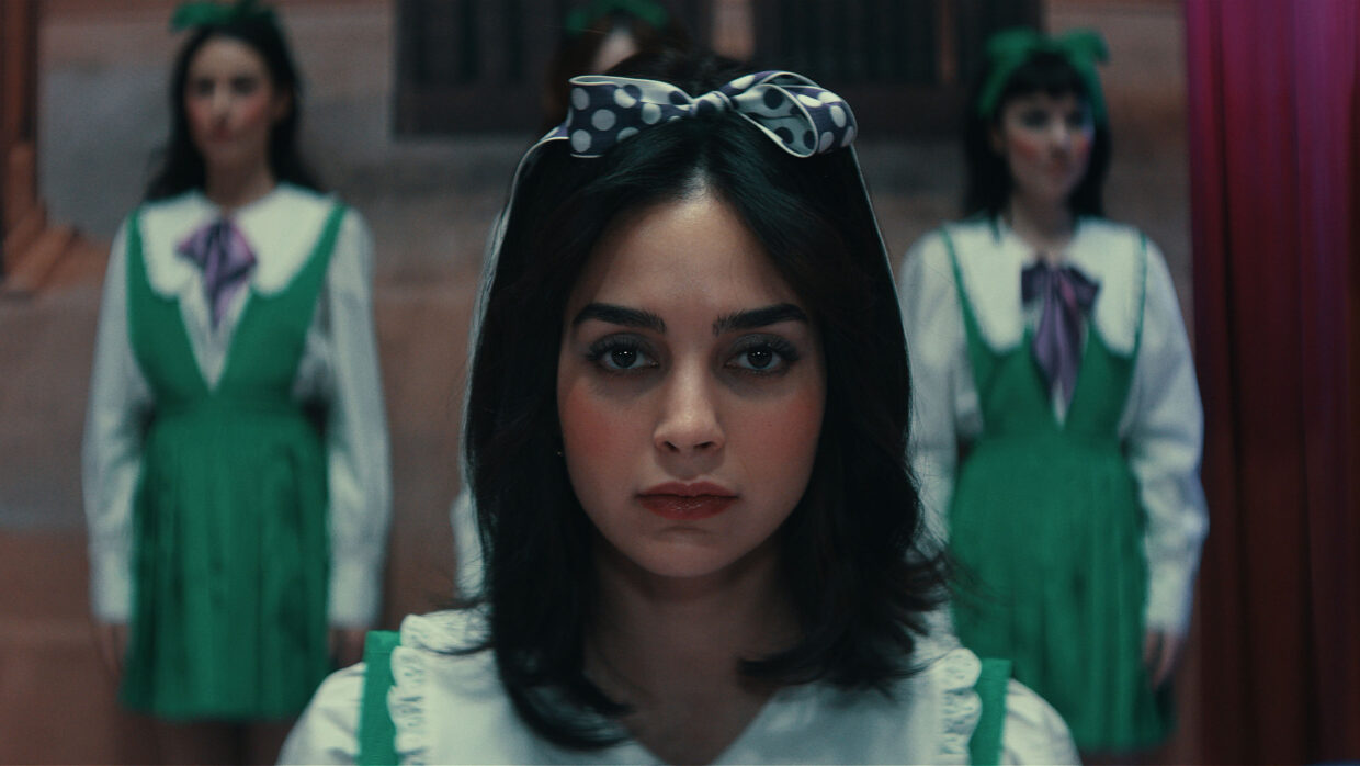 Three women with black hair dressed in green and white face toward the camera, one in close-up and two in the background.