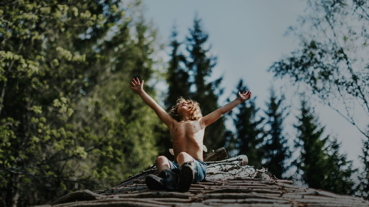 A blond, shirtless young boy sits in a forest, arms outstretched toward the sun.