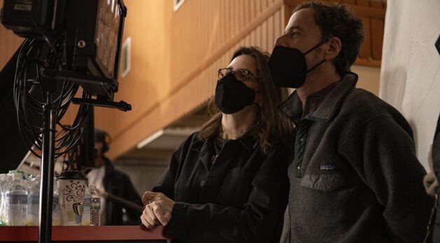 A woman and man, both wearing face masks, watch a monitor on-set.