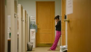 A woman in a black top and pink pants holds her face in despair in a deserted hallway.