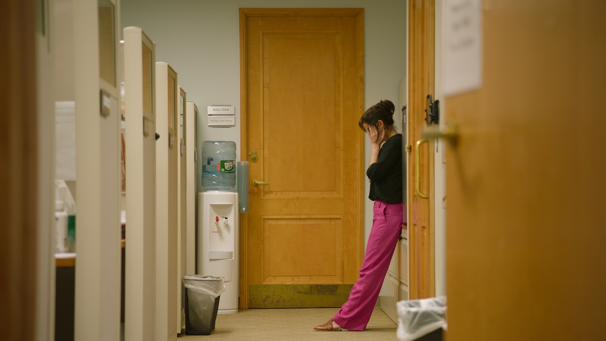 A woman in a black top and pink pants holds her face in despair in a deserted hallway.