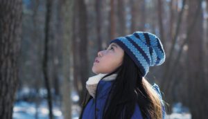 A young Japanese girl in a knit beanie stands staring upwards in a sunny forest during winter.
