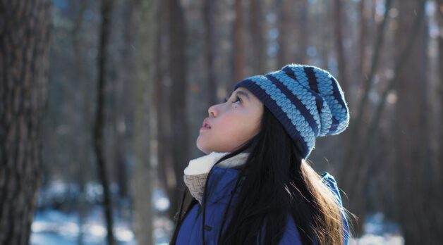 A young Japanese girl in a knit beanie stands staring upwards in a sunny forest during winter.